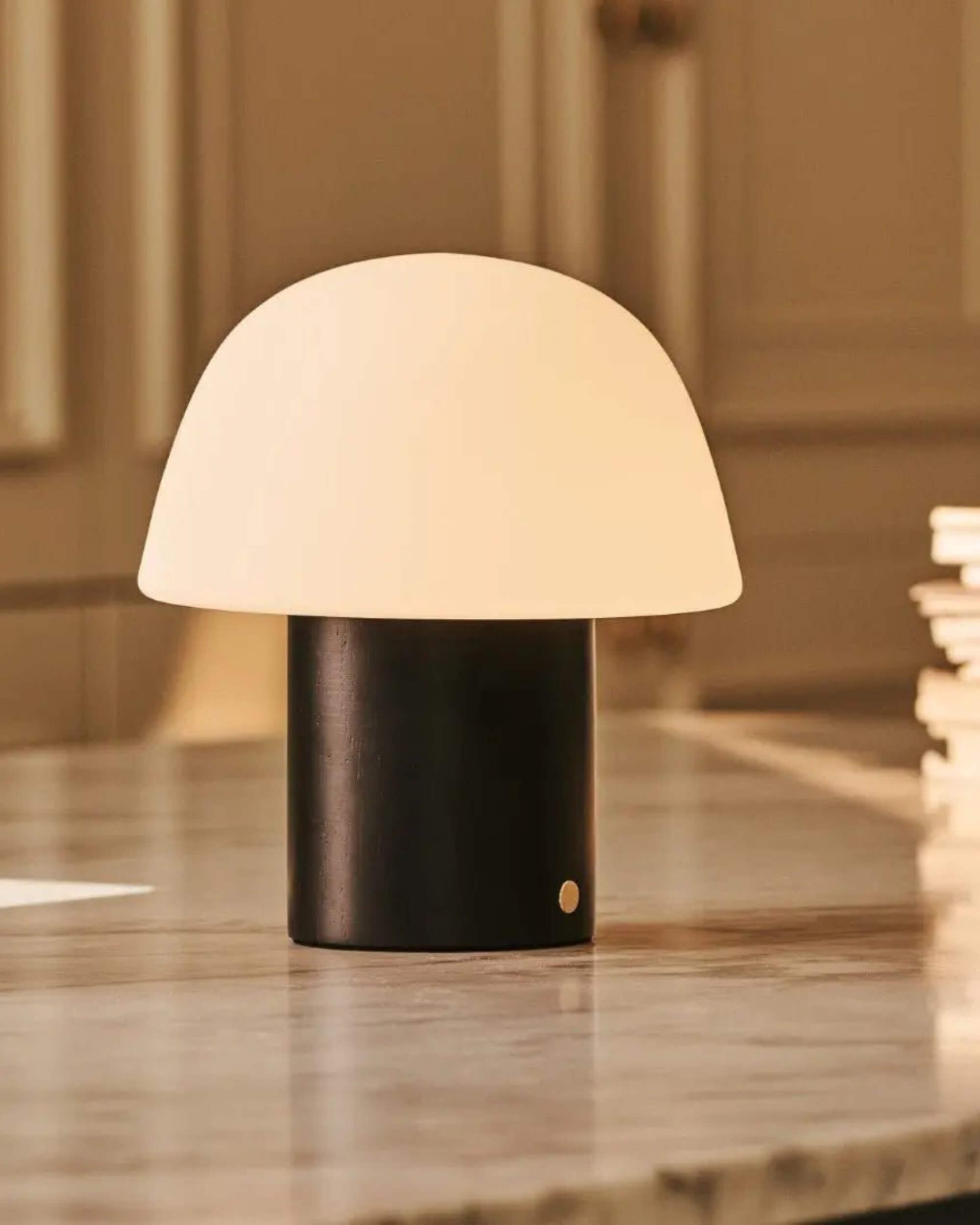 Lampe à poser nomade moderne avec pied cylindrique noir et abat-jour dôme blanc, allumée sur une surface en marbre. Bouton variateur tactile doré visible sur la base. Blomstra Home
