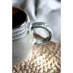 Gros plan sur l'anse ronde et l'émaillage réactif texturé de la tasse.