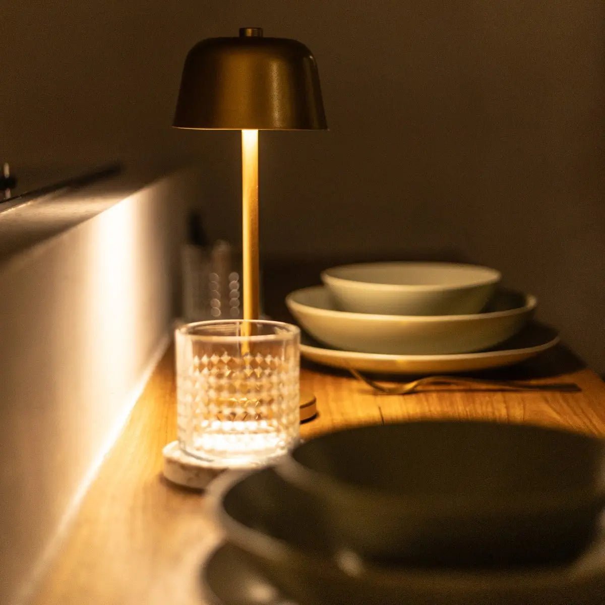 Lampe design minimaliste dorée posée sur une étagère comme objet de décoration, avec des assiettes et des couverts.