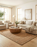 Grand tapis rectangulaire en jute tressé naturel Elliot Bronze (160x230 cm) disposé dans un salon spacieux et lumineux de style bohème chic. Il est placé sous un grand canapé d'angle beige, des fauteuils assortis et une table basse ronde en bois massif, sur un parquet clair.