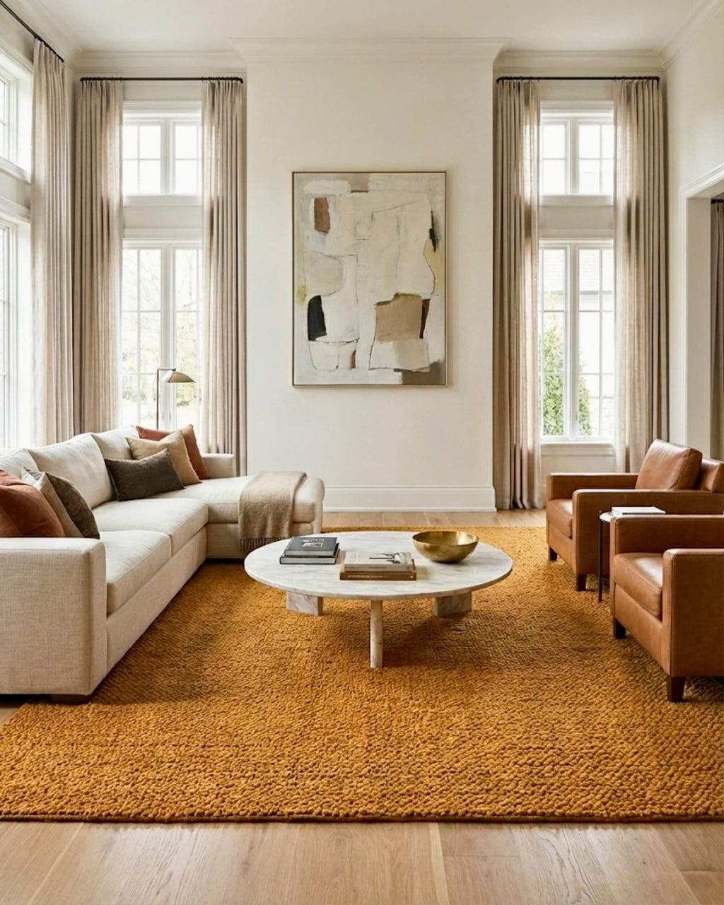 Très grand tapis rectangulaire en jute naturel tressé couleur moutarde Elios dans un salon spacieux et luxueux baigné de lumière naturelle. Il est placé sous un grand canapé d'angle beige, deux fauteuils en cuir marron et une table basse ronde en marbre blanc, devant de hautes fenêtres.
