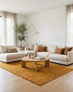 Grand tapis rectangulaire tressé en jute naturel couleur moutarde Elios (format 160x230) dans un salon moderne et lumineux. Il est placé au centre sur un parquet clair, sous un grand canapé d'angle beige avec des coussins ocre et une table basse ronde en bois. Un grand lampadaire arc est visible en arrière-plan.