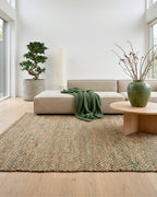 Grand tapis rectangulaire en jute et coton tressé Elliot Fougère (160x230 cm) disposé dans un salon lumineux au style scandinave et zen. Il est placé sur un parquet clair, sous une table basse ronde en bois et un grand canapé d'angle beige agrémenté d'un plaid vert. Un grand bonsaï, un vase vert avec des branches et un lampadaire en papier complètent cette décoration naturelle.