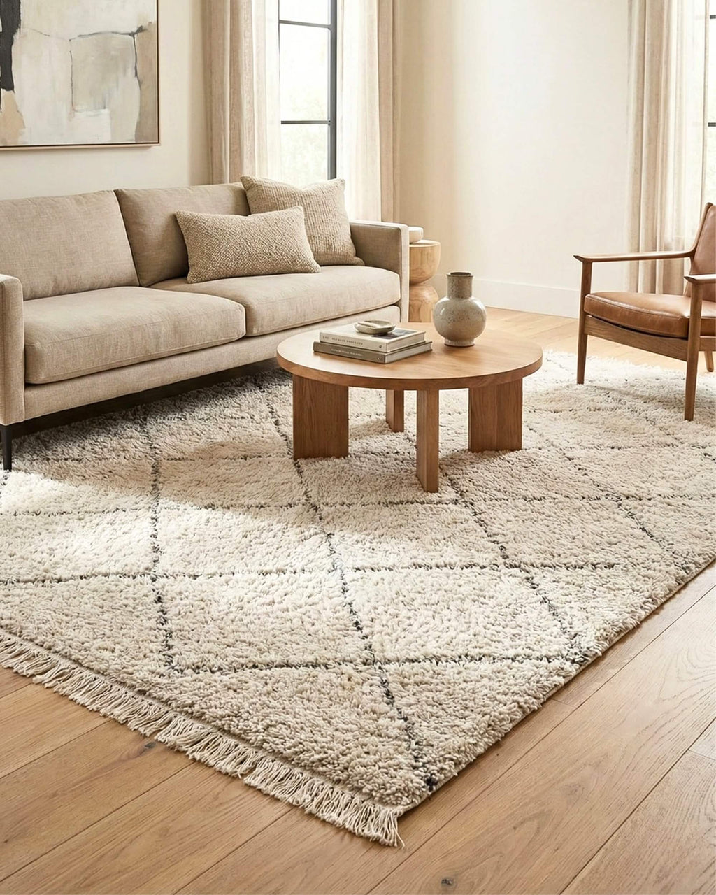 Grand tapis de salon style berbère Berry Neige, blanc cassé à motifs losanges gris fins et franges. Il est disposé dans un salon scandinave lumineux sous une table basse ronde en bois, accompagné d'un canapé beige et d'un fauteuil en cuir, créant une atmosphère chaleureuse.