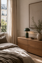 Lampe champignon fer texturé vert sauge allumée posée sur commode bois chêne clair dans chambre de style haussmannien, vase grès beige avec rameau d'olivier à droite, petit bol céramique sombre devant, rideau lin beige drapé et fenêtre à balcon ferronnerie noire avec vue sur immeubles parisiens, tableau minimaliste cadre bois au-dessus, lit linge stonewash taupe en avant-plan, ambiance chambre parisienne épurée et chaleureuse

