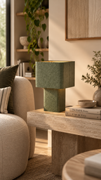 Lampe fer martelé vert kaki allumée avec intérieur doré rayonnant, posée sur console travertin beige avec livres The Kinfolk Home et L'Art de Vivre, vase céramique blanc sphérique avec branches vertes à droite, petit bol céramique beige, fauteuil bouclette beige arrondi à gauche avec coussins kaki et rayures, étagère bois avec plante monstera et vase céramique blanc en arrière-plan, tableau bois sable encadré, lumière naturelle dorée, ambiance salon nordique lumineux