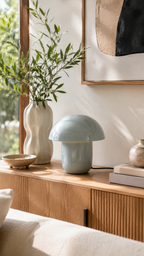 Lampe NORA champignon bleu céladon posée sur buffet cannelé bois chêne, grand vase céramique blanc ondulé avec branches d'olivier à gauche, bol céramique beige, livres empilés, tableau abstrait encadré bois noir et beige en arrière-plan, baie vitrée avec lumière naturelle, ambiance salon contemporain nordique fond beige