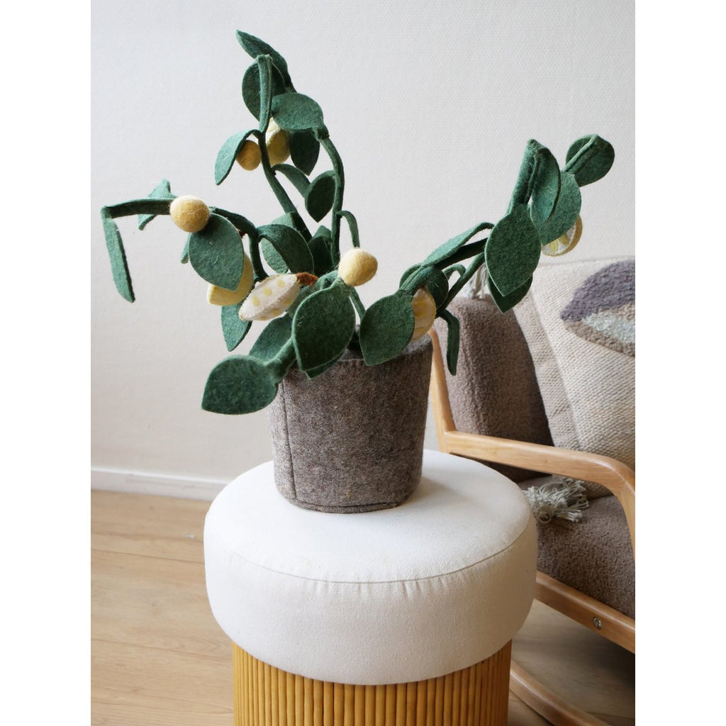 Plante décorative citronnier en feutre vert et jaune posée sur un pouf cylindrique moutarde habillé de toile blanche, parquet bois clair, fauteuil bois naturel avec plaid tufté gris-lilas et coussin rond en arrière-plan à droite, mur blanc lumineux

