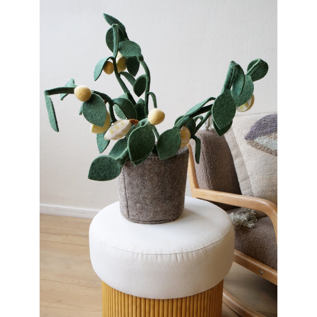 Plante décorative citronnier en feutre vert et jaune posée sur un pouf cylindrique moutarde habillé de toile blanche, parquet bois clair, fauteuil bois naturel avec plaid tufté gris-lilas et coussin rond en arrière-plan à droite, mur blanc lumineux

