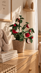 Plante décorative pommier en feutre pommes rouges posée au centre d'une commode cannage rotin bois clair dans une chambre bébé lumineuse, rideau voile blanc à droite, peluche lapin beige et livres empilés à gauche, affiche encadrée "petit amour ♥" au mur en arrière-plan