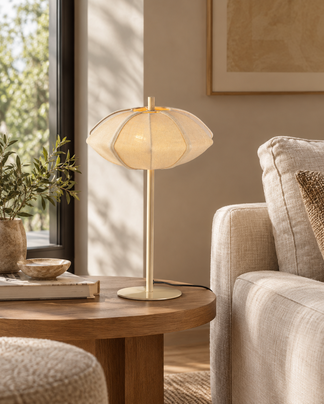 Lampe de table PARIS Disque, abat-jour en coton beige crème et pied doré, sur une table en bois près d’un canapé écru.
