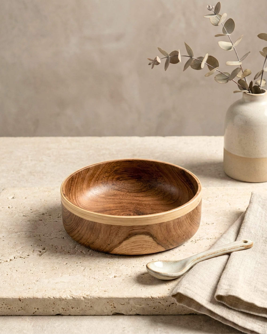 Bol artisanal Soraya en bois de teck avec une bordure contrastée en bambou clair. Posé sur une table en travertin avec une cuillère en céramique.