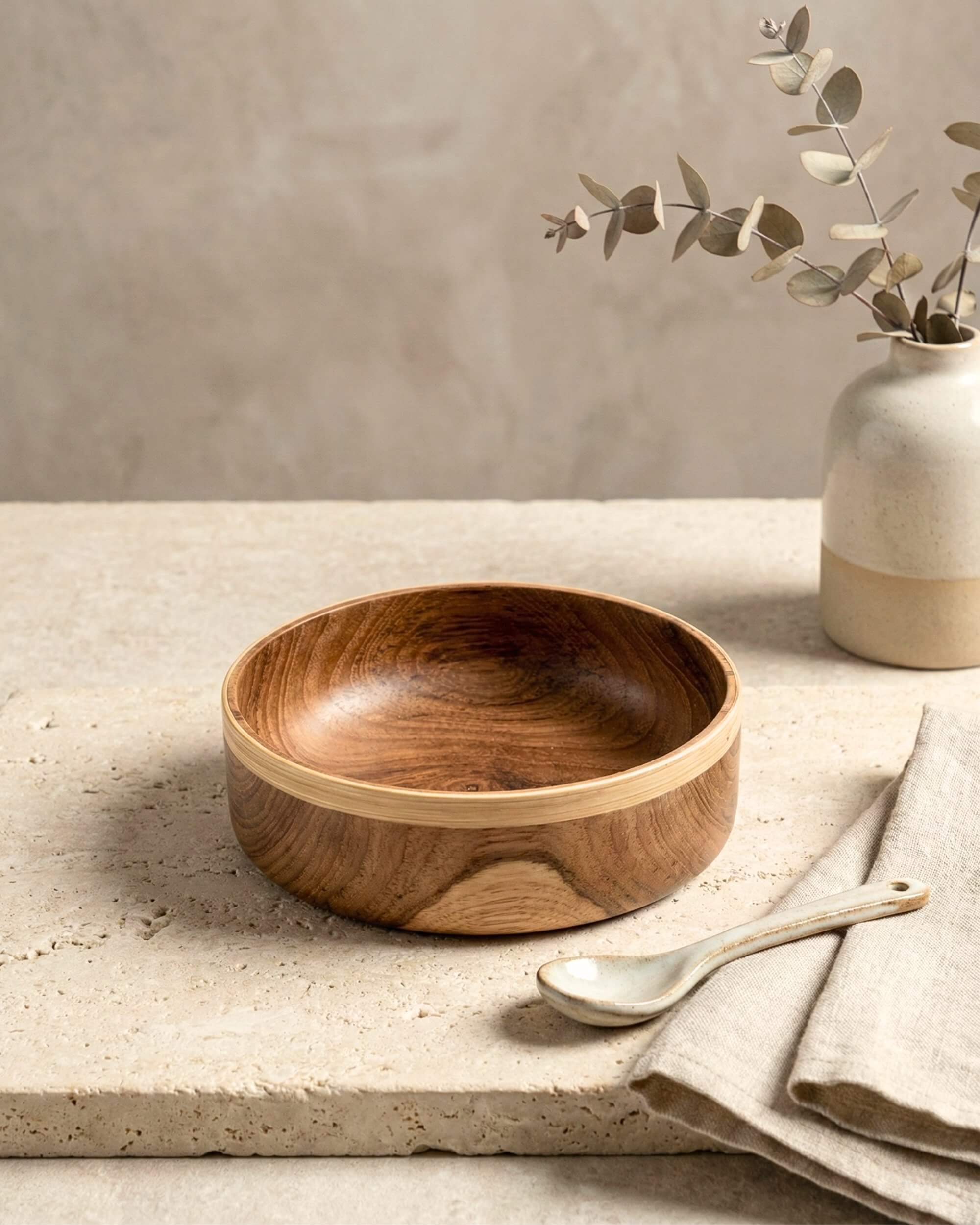 Bol artisanal Soraya en bois de teck avec une bordure contrastée en bambou clair. Posé sur une table en travertin avec une cuillère en céramique.