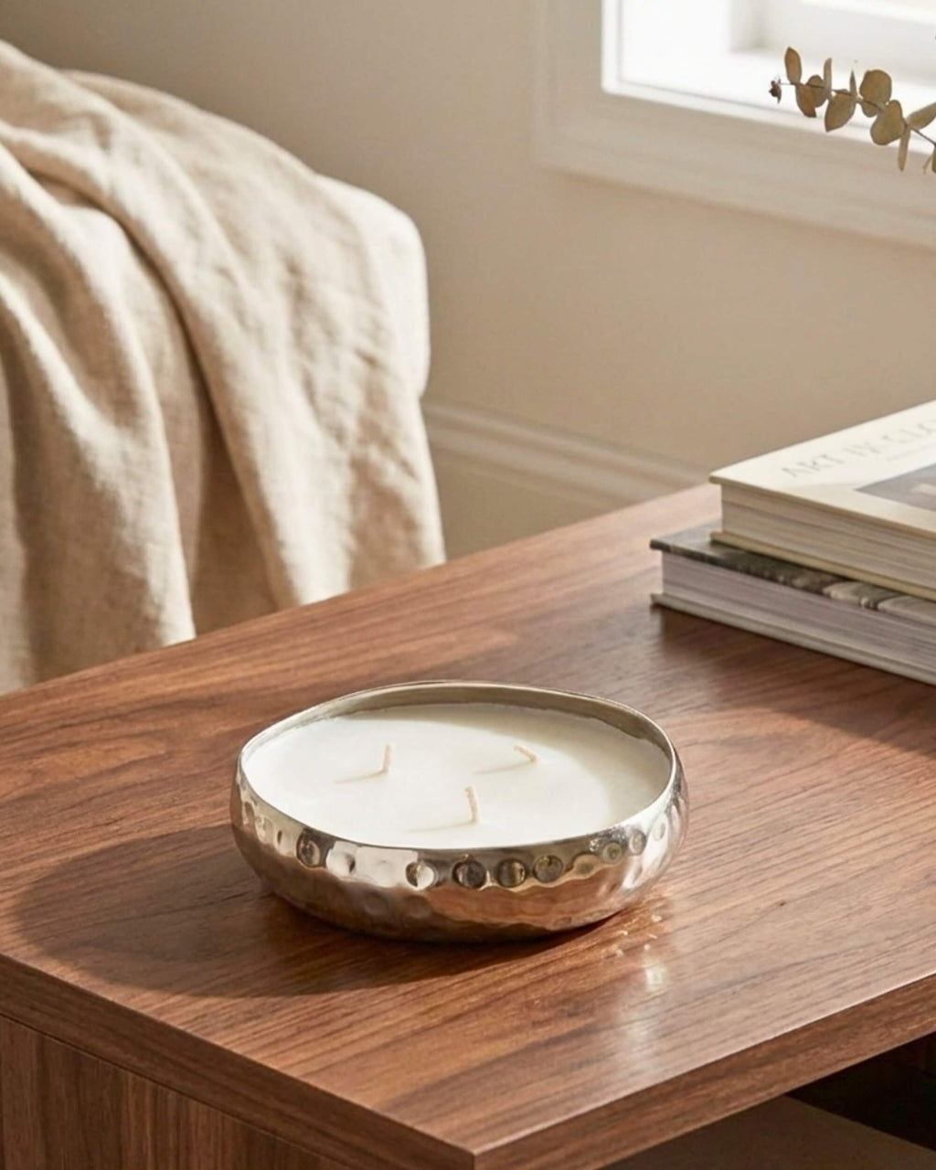 Bougie parfumée 3 mèches dans sa coupe en métal argenté martelé, posée sur une table basse en bois dans un salon moderne.
