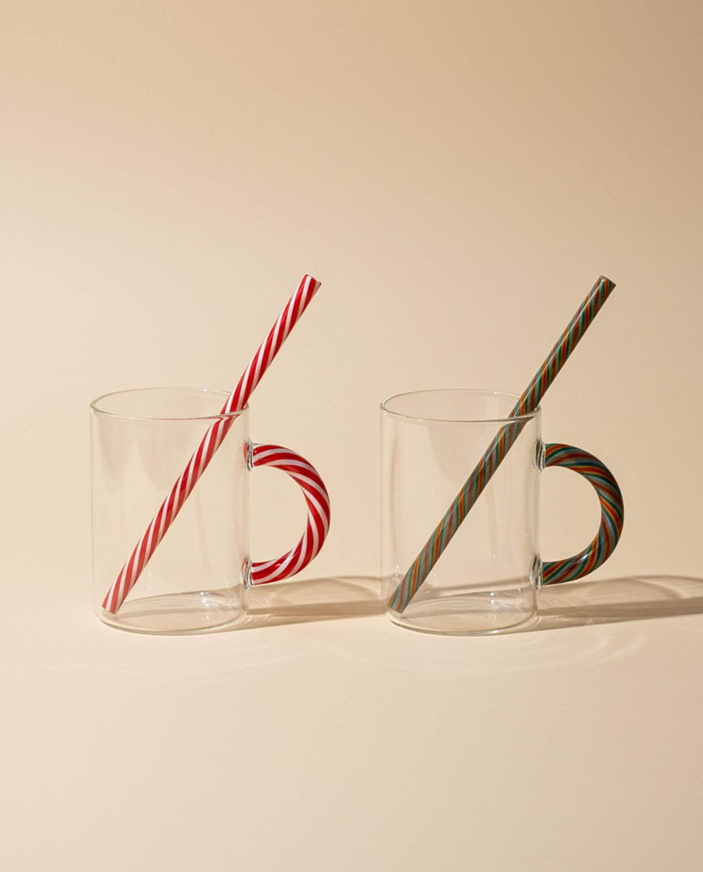 Lot de deux tasses CANDY CANE en verre soufflé avec anse rayée rouge et verte sur fond beige.
