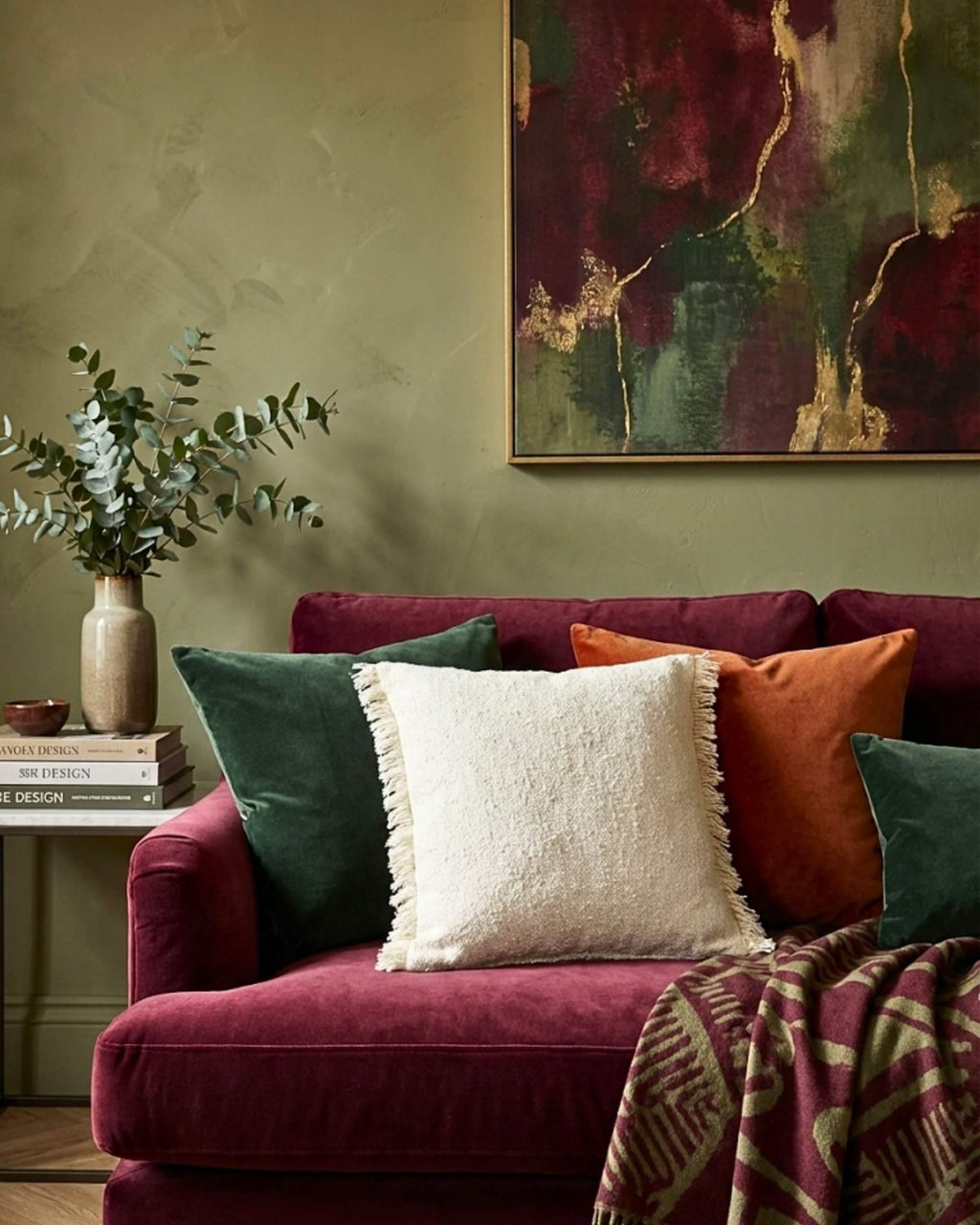 Coussin carré en tissu bouclette écru et franges, mis en scène sur un canapé en velours bordeaux dans un salon cosy avec mur vert et décoration murale abstrait.