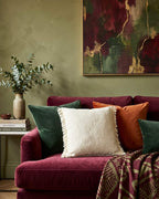 Coussin carré en tissu bouclette écru et franges, mis en scène sur un canapé en velours bordeaux dans un salon cosy avec mur vert et décoration murale abstrait.