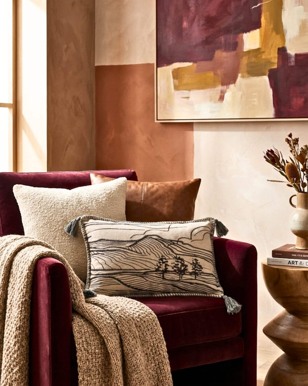 Mise en scène salon automnal : coussin lombaire brodé paysage 'Vallée' posé sur un fauteuil en velours bordeaux, accompagné d'un plaid en grosse maille et d'objets déco sur une table en bois sculpté.