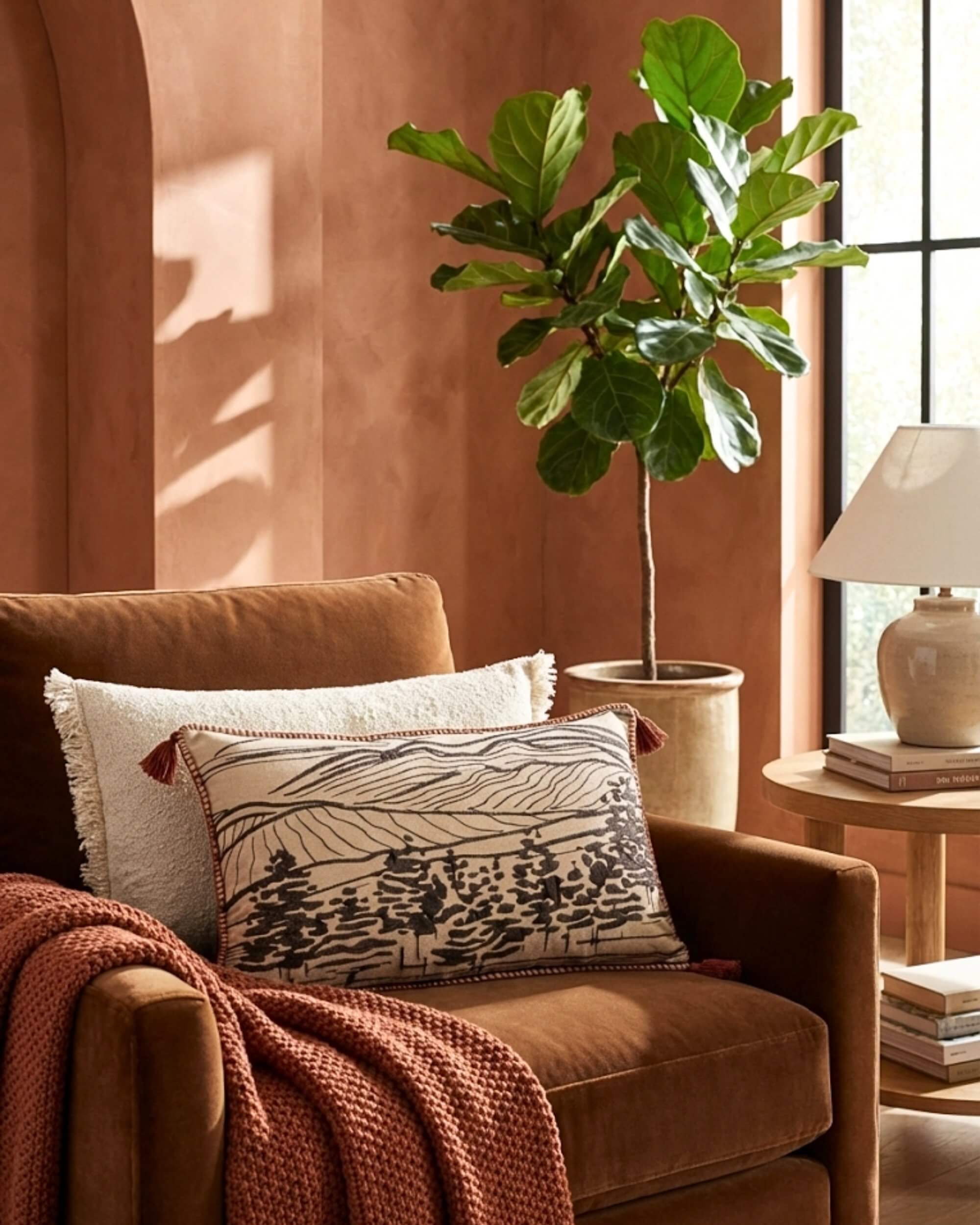 Grand coussin lombaire brodé Vallée couleur sienne posé sur un fauteuil en velours marron, accompagné d'un plaid en maille rouille et d'une grande plante verte.