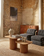 Ambiance salon loft : duo de tables basses organiques en bois d'acacia massif, disposées sur un tapis beige face à des fauteuils gris dans une pièce aux murs bruts.