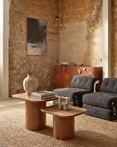 Ambiance salon loft : duo de tables basses organiques en bois d'acacia massif, disposées sur un tapis beige face à des fauteuils gris dans une pièce aux murs bruts.