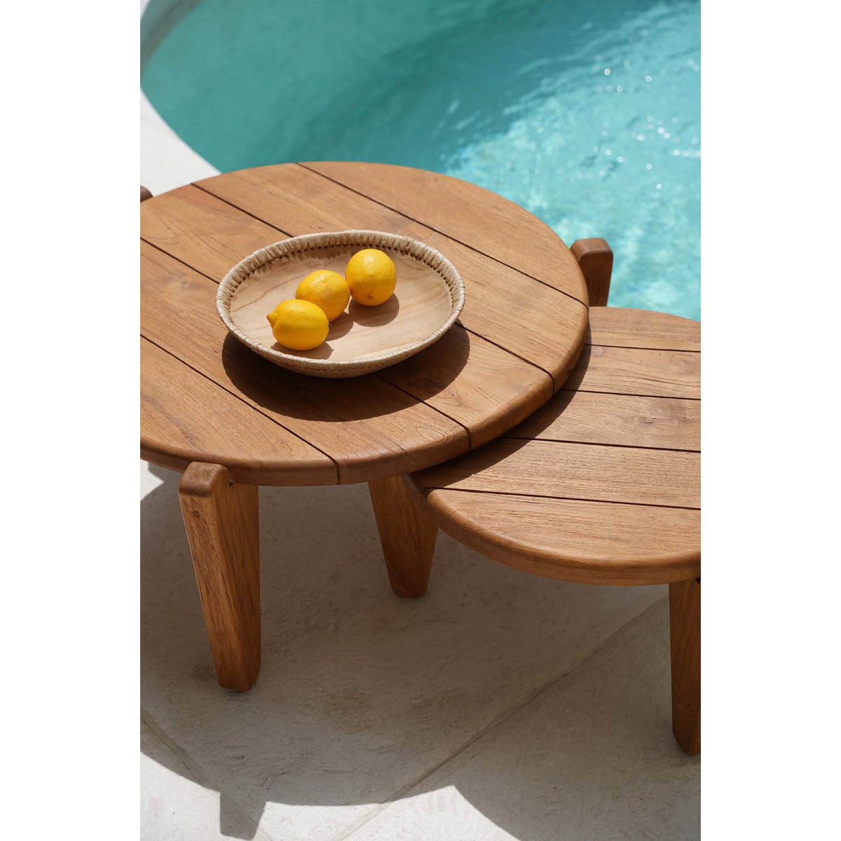 Mise en scène estivale de la table basse en teck posée au bord d'une piscine.