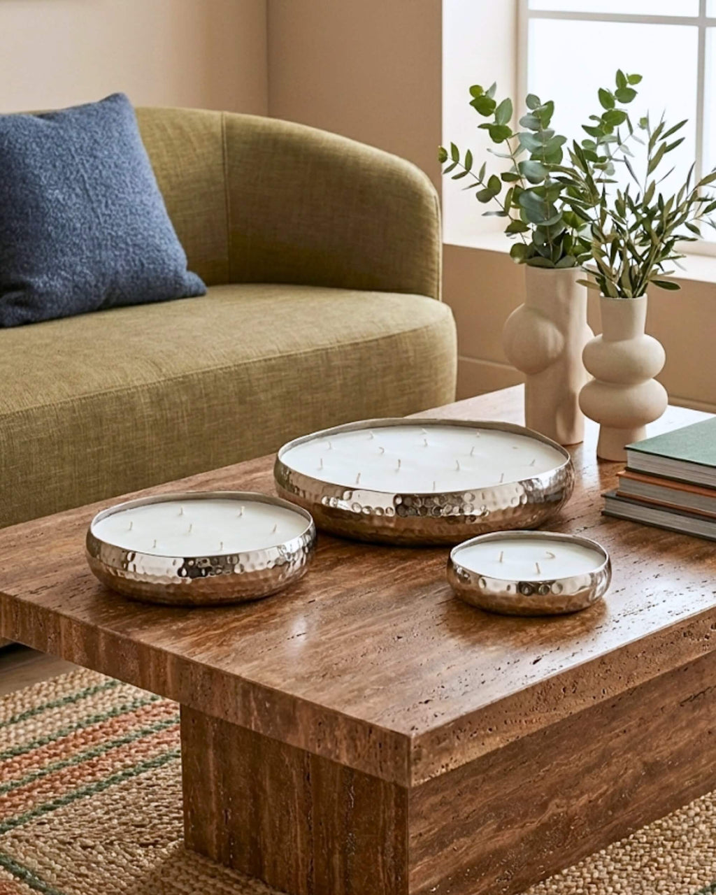Idée décoration : trio de bougies multi-mèches en coupes de métal argenté martelé, disposées sur une table en bois avec vases et feuillage d'eucalyptus.