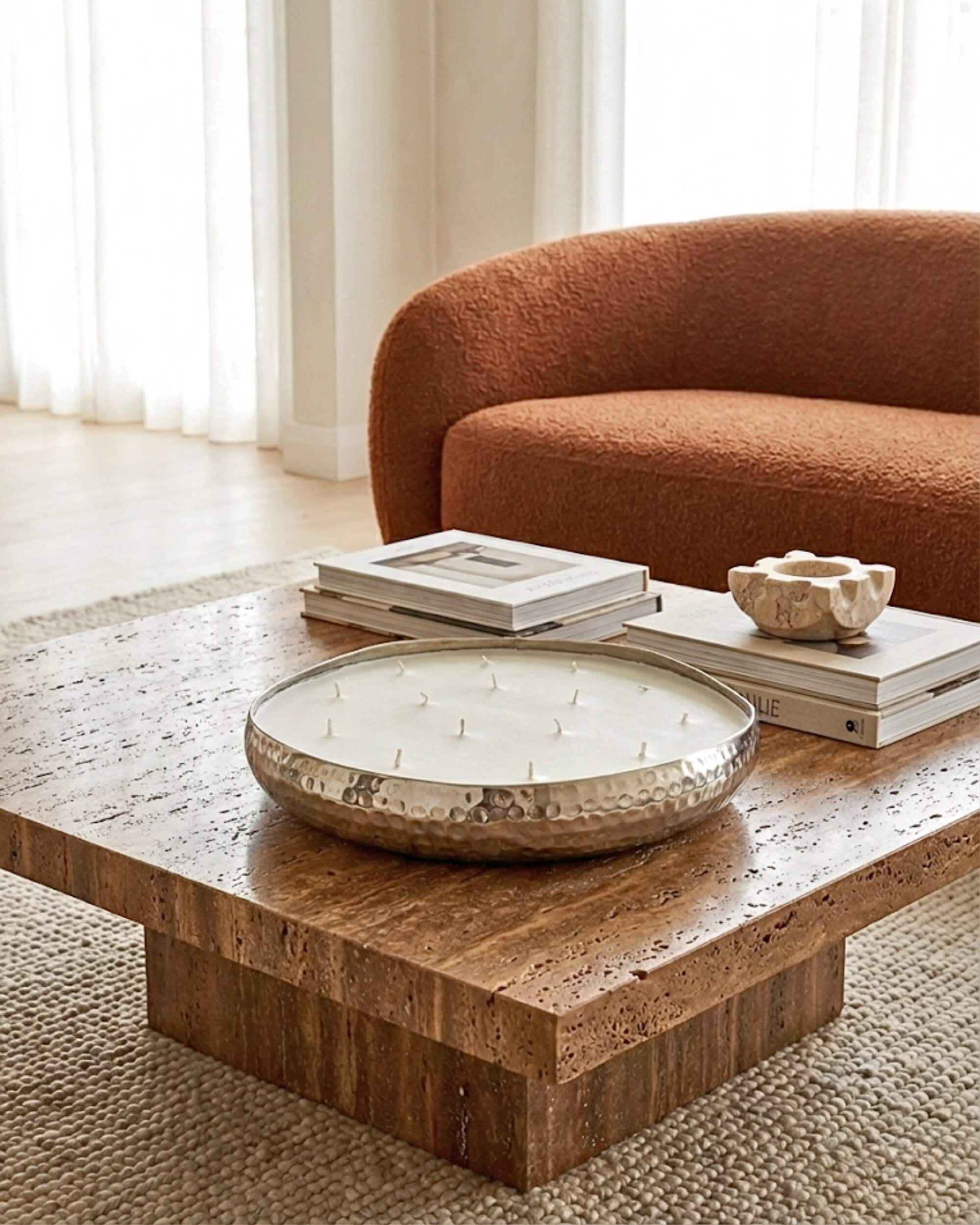 Grande bougie centre de table multi-mèches dans une coupe en métal argenté martelé, posée sur une table basse en travertin dans un salon à la décoration chaleureuse avec un canapé terracotta.