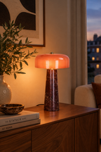 Lampe LAZIO Cône Grand allumée sur un meuble en bois, près d’une plante, avec un abat-jour aux reflets orangés.