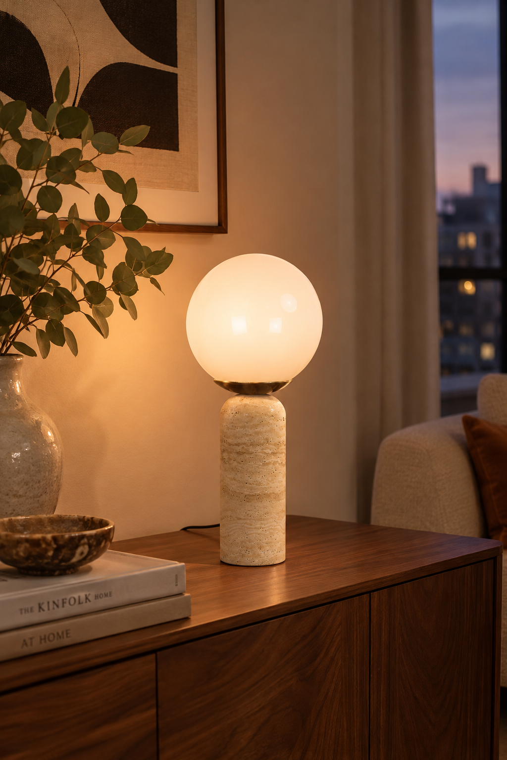 Lampe LAZIO Sphère crème allumée sur un meuble en bois, près de livres et d’une plante.
