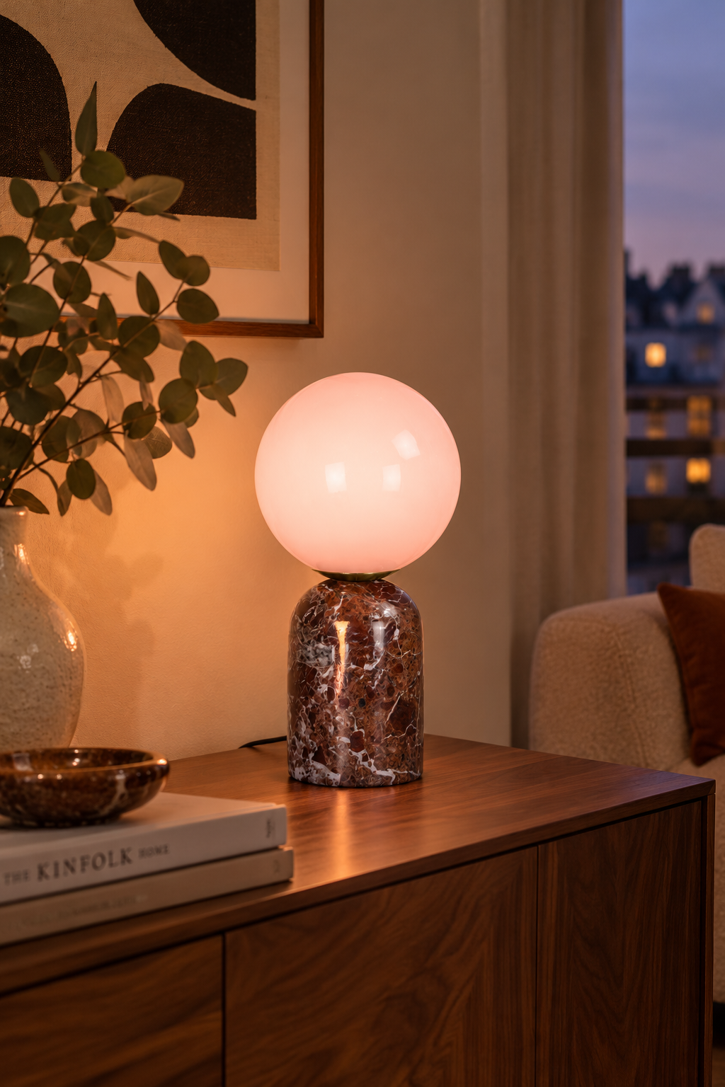 Petite lampe LAZIO Sphère allumée sur un meuble en bois, avec un globe lumineux aux nuances rosées.