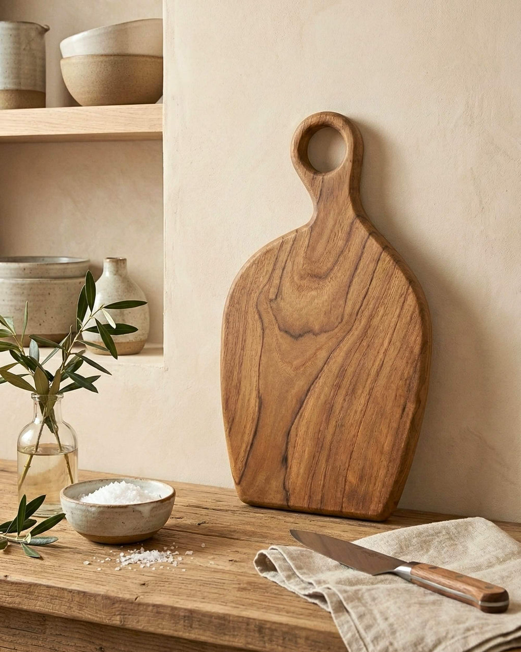 Planche à découper artisanale Duma en bois massif avec poignée sculptée, posée à la verticale. Ambiance cuisine rustique chic avec sel de mer et branche d'olivier.