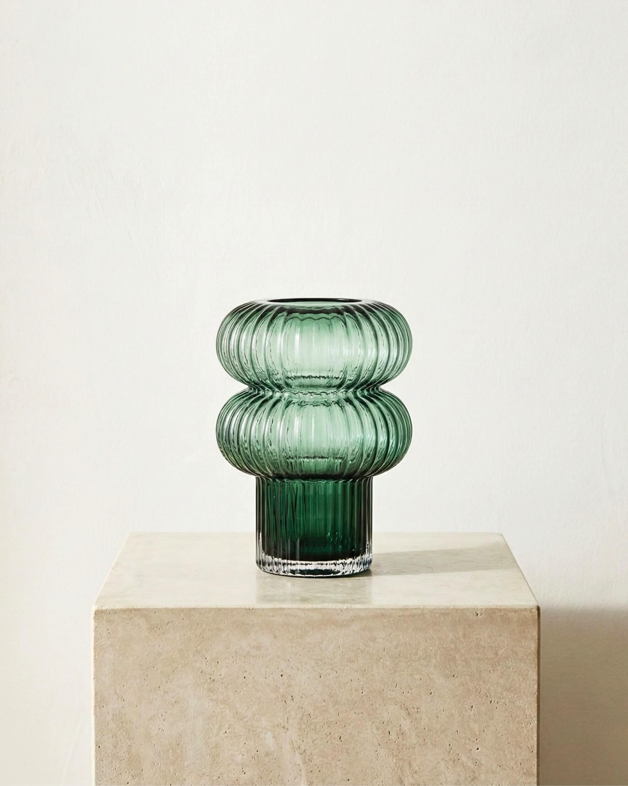 Vase design Aurora en verre cannelé vert émeraude. Forme sculpturale à double bulle sur pied, posé sur un socle en travertin beige.