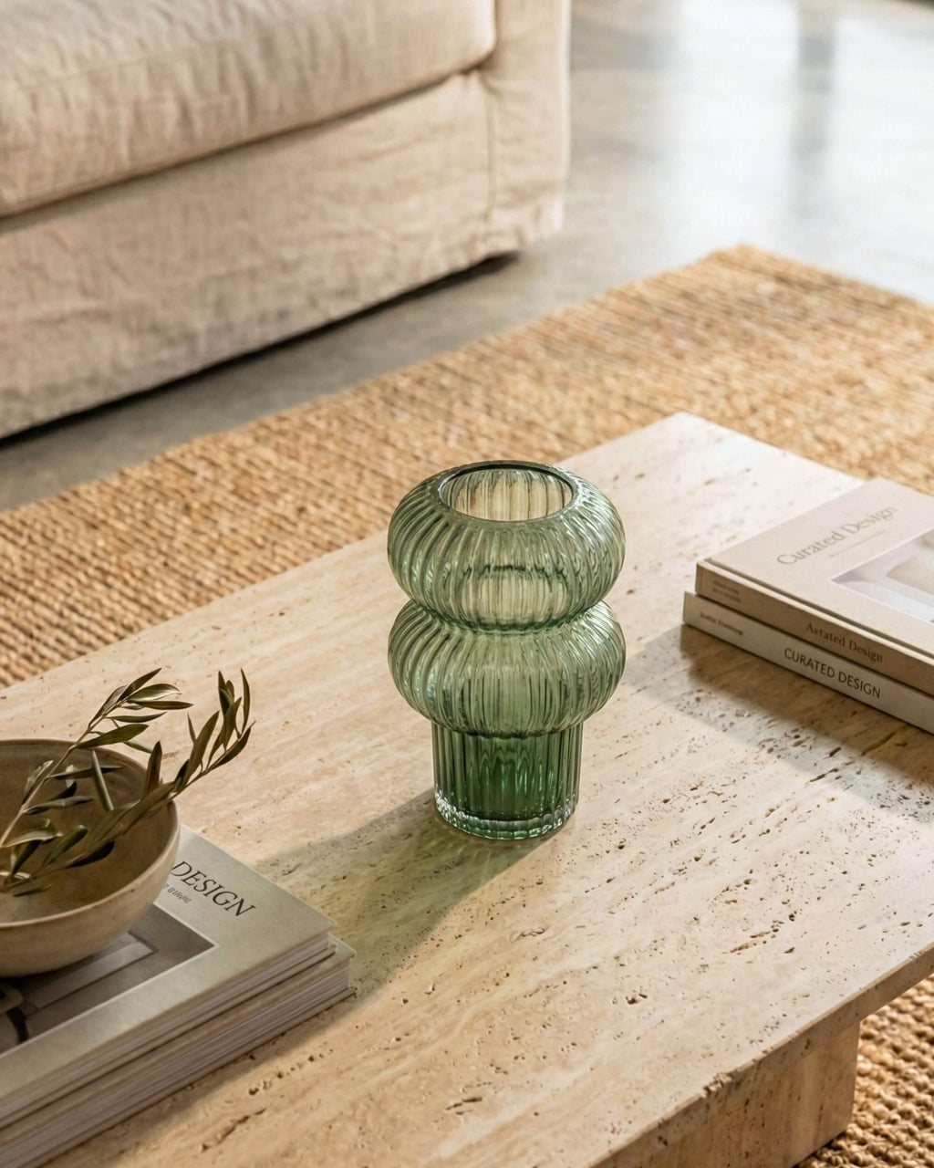 Vase Aurora en verre cannelé vert émeraude mis en scène sur une table basse en travertin. Ambiance salon cosy avec livres de design et canapé beige.
