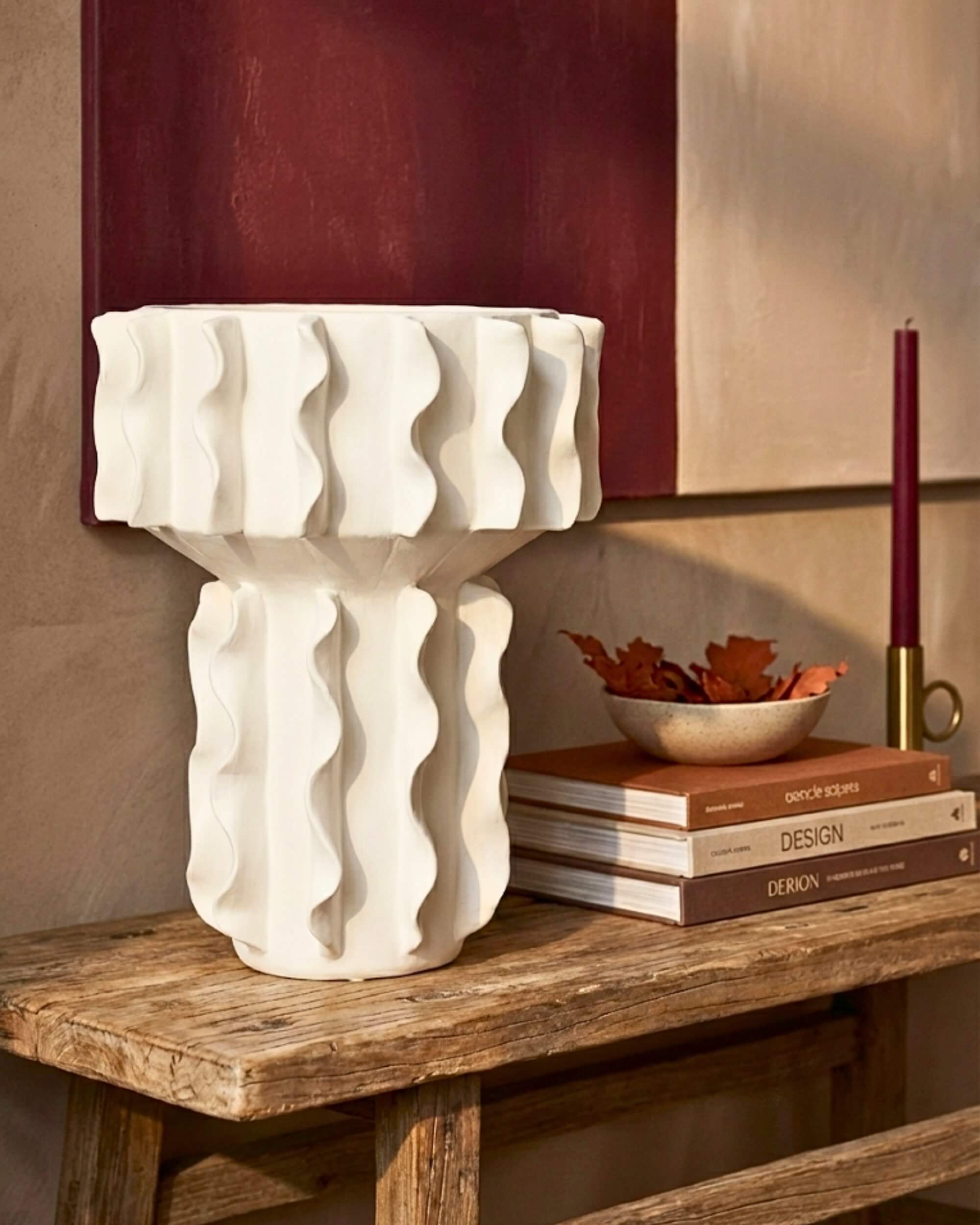 Gros plan sur le vase sculptural Hélios en céramique blanche ondulée, posé sur une console en bois brut avec des livres déco et un bougeoir.
